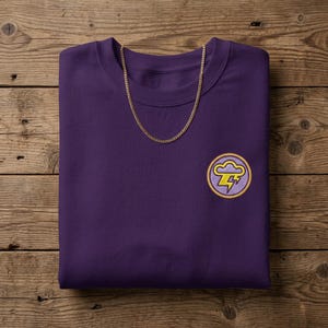 Peut inclure: Un sweat-shirt à col rond violet foncé avec un petit écusson brodé circulaire sur la poitrine gauche. L'écusson représente un nuage violet clair avec un éclair jaune à l'intérieur. Une fine chaîne en métal doré repose au niveau du col.