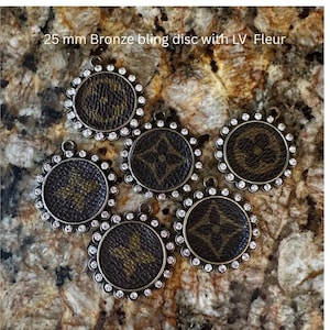Puede incluir: Cinco dijes circulares de bronce de 25 mm con un material marrón estampado con un diseño repetitivo de flores y monogramas. Cada dije está bordeado por un anillo de pequeños cristales transparentes e incluye una pequeña argolla.