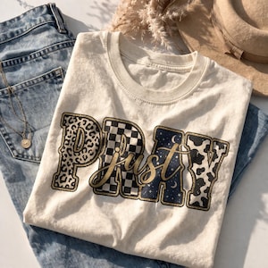 Könnte beinhalten: Cremefarbenes T-Shirt mit dem Schriftzug "Just Pray" in dekorativer Schrift. Die Buchstaben haben verschiedene Muster, darunter Leopardenmuster, ein Schachbrettmuster und einen Sternenhimmel. Das Shirt liegt auf einer blauen Jeans.