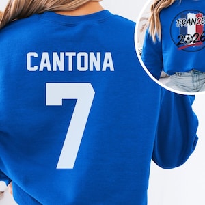 Blue retro soccer tee featuring CANTONA 7 printed in solid white on the back. A small inset circle displays the front design containing the French flag colors, a football, and the year 2026
