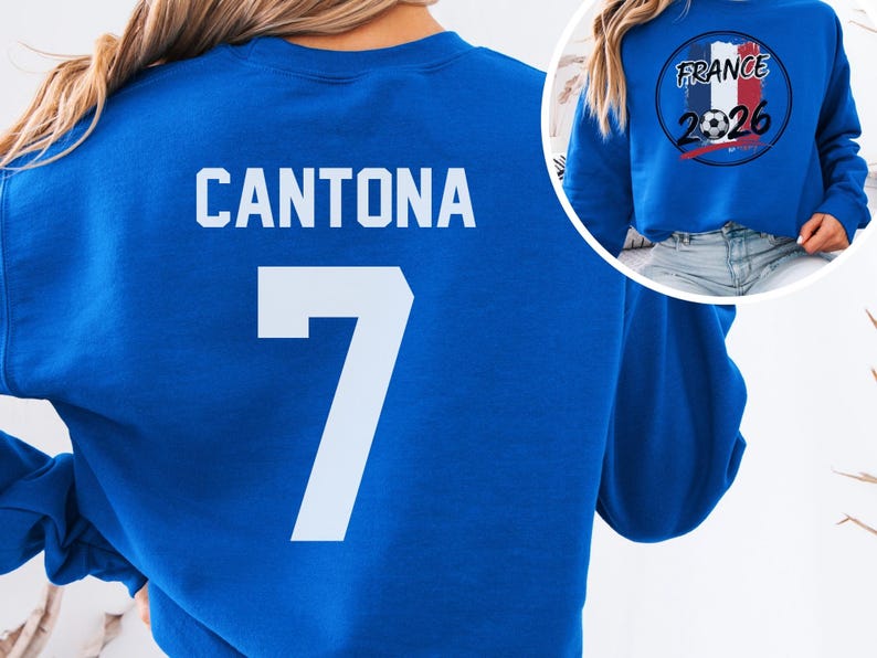 Blue retro soccer tee featuring CANTONA 7 printed in solid white on the back. A small inset circle displays the front design containing the French flag colors, a football, and the year 2026