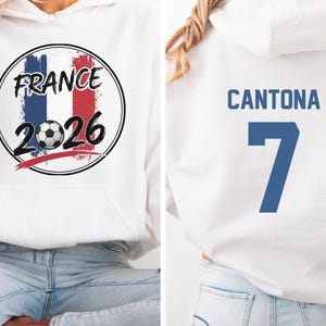 Puede incluir: Sudadera con capucha blanca con un gr&aacute;fico frontal con la palabra "FRANCE" encima de un bal&oacute;n de f&uacute;tbol y el a&ntilde;o "2026" en negro. La parte trasera de la sudadera muestra el nombre "CANTONA" y el n&uacute;mero "7" en azul.