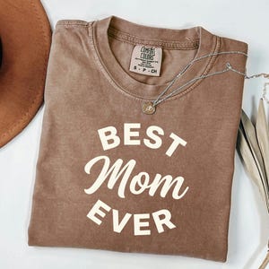 May include: A brown Comfort Colors t-shirt with the text "BEST Mom EVER" in white. A silver necklace with a pendant is draped across the shirt. A brown hat and dried palm leaves are in the background.