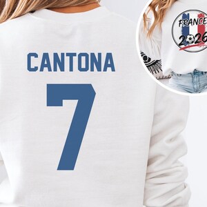 White short-sleeve t-shirt featuring CANTONA 7 printed in classic blue on the back. A small inset circle shows the front graphic with a vintage France flag, a soccer ball, and the text FRANCE 2026.