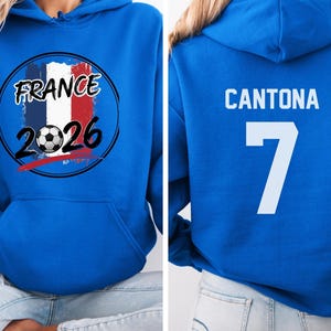 Puede incluir: Una sudadera con capucha azul real con un gr&aacute;fico de la bandera francesa, un bal&oacute;n de f&uacute;tbol y el texto "FRANCE 2026". La parte trasera de la sudadera muestra el nombre "CANTONA" y el n&uacute;mero "7" en blanco. La sudadera tiene un bolsillo delantero y una capucha.