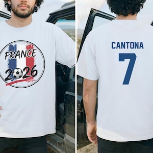 Puede incluir: Camiseta blanca con un gr&aacute;fico en la parte delantera que presenta los colores de la bandera francesa, un bal&oacute;n de f&uacute;tbol y el texto "FRANCE 2026". La parte trasera de la camiseta muestra el nombre "CANTONA" y el n&uacute;mero "7" en azul.