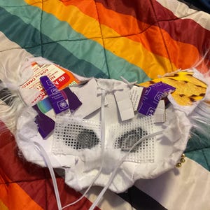 May include: An unfinished white mask with black eye openings and a white elastic strap. The mask features a mesh-like material over the eye openings. The mask rests on a rainbow-striped blanket.