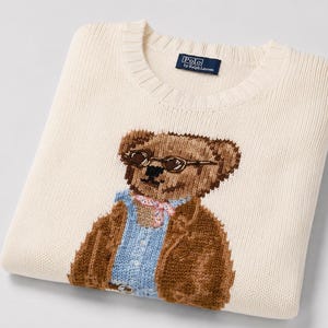 May include: A cream-colored crewneck sweater featuring a knit graphic of a brown teddy bear wearing round glasses, a light blue button-down shirt, and a patterned neckerchief. The interior collar tag displays the text Polo by Ralph Lauren.