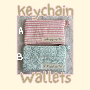 May include: Two rectangular fabric keychain wallets with zippers and side tabs. Wallet A features pink and white horizontal stripes. Wallet B has a green and white floral pattern. Both have a white label reading "peony projects".