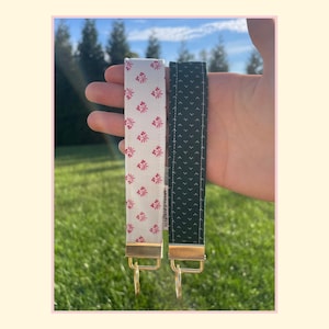 May include: Two fabric wristlet keychains with gold-tone metal hardware. One strap is white with a repeating red floral pattern, and the other strap is black with a repeating white dot pattern. Both feature a small white fabric tag on the side.