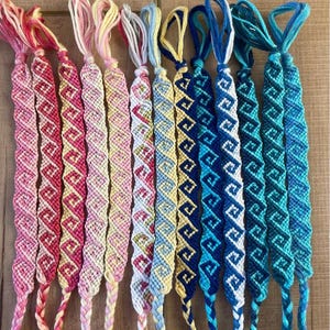 May include: A collection of twelve woven friendship bracelets arranged in a row, featuring a repeating geometric wave pattern. The bracelets display a gradient of colors ranging from warm pinks and yellows to cool blues and teals, each with braided ends.