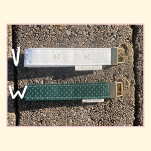 May include: Two fabric wristlet keychains with gold-tone hardware. The top keychain is white with a small floral pattern. The bottom keychain is dark green with a white dot pattern. Both feature a small white tag labeled peony projects.