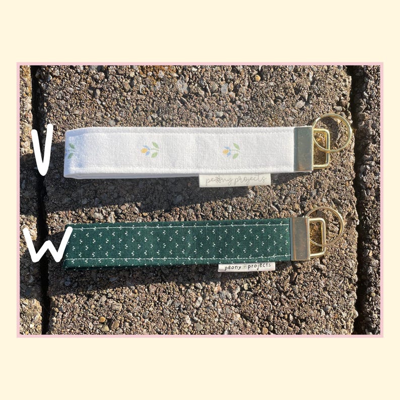 May include: Two fabric wristlet keychains with gold-tone hardware. The top keychain is white with a small floral pattern. The bottom keychain is dark green with a white dot pattern. Both feature a small white tag labeled peony projects.
