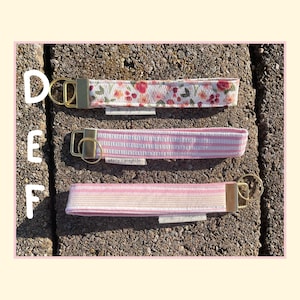 May include: Three fabric wristlet keychains labeled D, E, and F. Keychain D features a white floral print. Keychain E has pink and white vertical stripes. Keychain F displays horizontal pink, white, and yellow stripes. Each has a gold metal clasp.