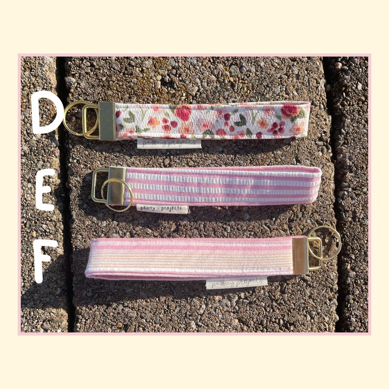 May include: Three fabric wristlet keychains labeled D, E, and F. Keychain D features a white floral print. Keychain E has pink and white vertical stripes. Keychain F displays horizontal pink, white, and yellow stripes. Each has a gold metal clasp.