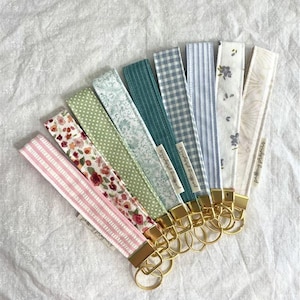 May include: A collection of fabric key fobs in various patterns and colors, including pink stripes, floral, polka dots, and blue gingham. Each fob has a gold-colored metal clasp and key ring.