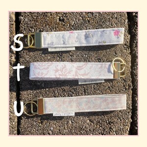 May include: Three fabric wristlet keychains with gold-toned metal hardware and split rings. Each strap features a different floral or damask pattern in soft pink, white, and grey tones. A small white tag labeled peony projects is attached to each.