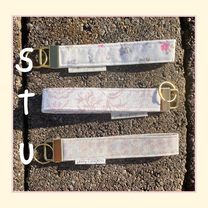 May include: Three fabric wristlet keychains with gold-toned metal hardware and split rings. Each strap features a different floral or damask pattern in soft pink, white, and grey tones. A small white tag labeled peony projects is attached to each.