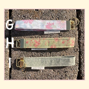 May include: Three fabric wristlet keychains labeled G, H, and I. G features a pink and blue floral pattern, H has a sage green floral print, and I is a solid grey textured fabric. Each includes a gold-tone metal clasp and a small Peony Projects tag.