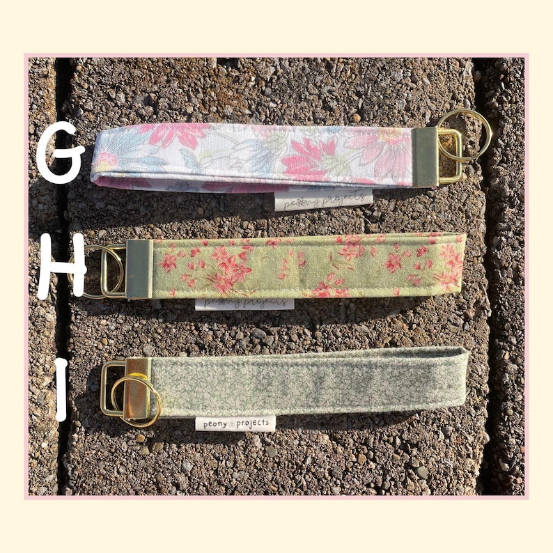 May include: Three fabric wristlet keychains labeled G, H, and I. G features a pink and blue floral pattern, H has a sage green floral print, and I is a solid grey textured fabric. Each includes a gold-tone metal clasp and a small Peony Projects tag.