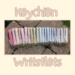 May include: A collection of fabric wristlet keychains featuring various patterns including red gingham, floral prints, stripes, and solid colors. Each keychain has a gold-toned metal clasp and ring. The text "keychain Writstlets" is visible.