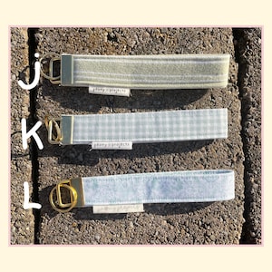May include: Three fabric wristlet keychains with gold-tone hardware and split rings. Top J is sage green with white stripes, middle K is light green gingham, and bottom L is light blue floral print. Each features a white Peony Projects label.