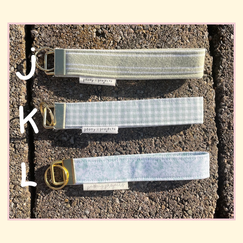 May include: Three fabric wristlet keychains with gold-tone hardware and split rings. Top J is sage green with white stripes, middle K is light green gingham, and bottom L is light blue floral print. Each features a white Peony Projects label.