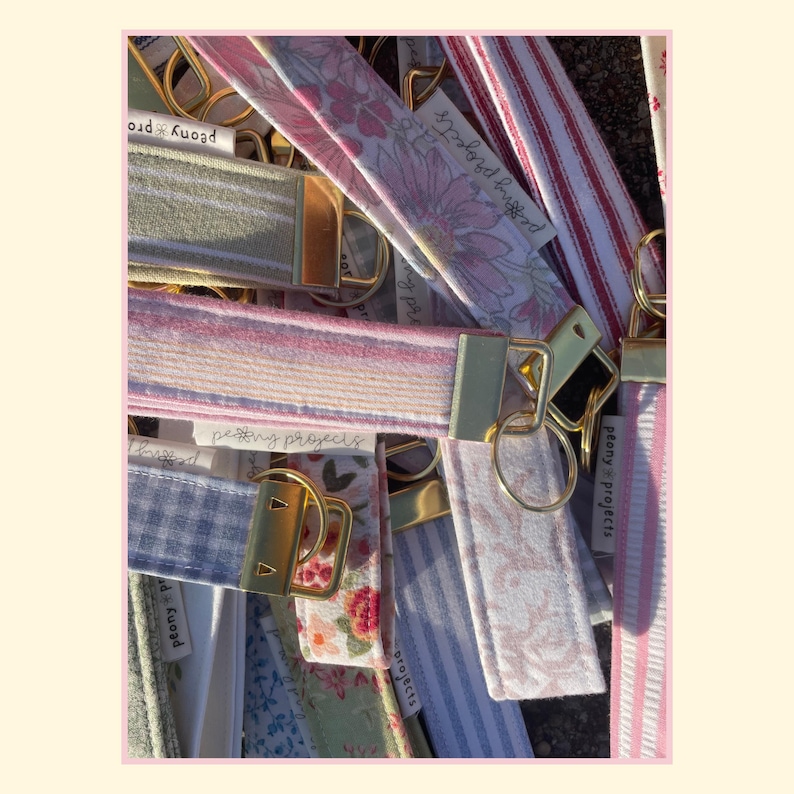 May include: A collection of fabric wristlet keychains featuring various patterns including floral prints, gingham checks, and stripes. Each strap is attached to a gold-toned metal clasp and a circular ring, with some labels reading Peony Projects.