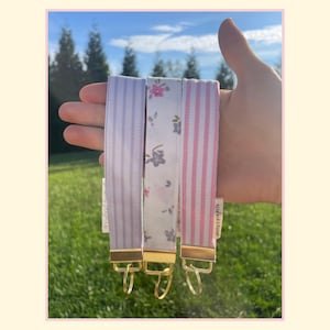 May include: Three fabric wristlet keychains with gold-tone hardware and split rings. One features light purple vertical stripes, one has a white background with a small pink and grey floral pattern, and one has light pink vertical stripes.