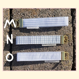 May include: Three fabric wristlet keychains featuring light blue and white patterns, including stripes and gingham checks. Each keychain has a gold-tone metal clasp and a split ring, with a small white tag labeled peony projects attached.