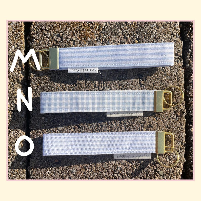 May include: Three fabric wristlet keychains featuring light blue and white patterns, including stripes and gingham checks. Each keychain has a gold-tone metal clasp and a split ring, with a small white tag labeled peony projects attached.