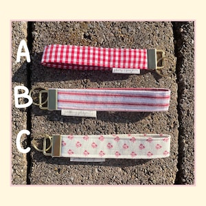 May include: Three fabric wristlet keychains with gold-tone hardware and split rings. Option A features a red and white gingham pattern. Option B has red and white vertical stripes. Option C displays a small red floral print on a cream background.
