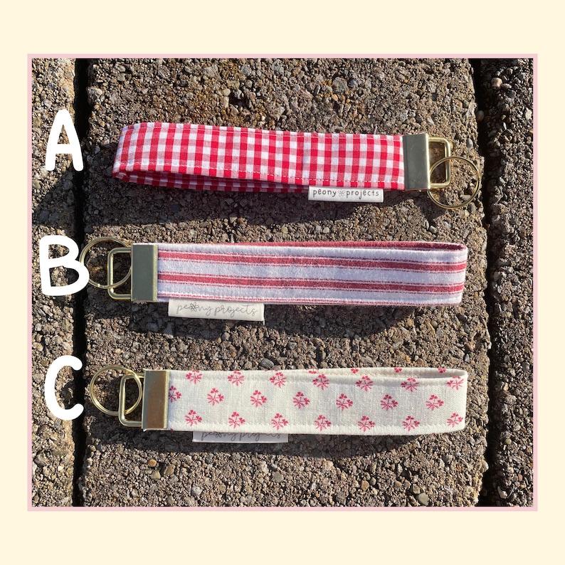 May include: Three fabric wristlet keychains with gold-tone hardware and split rings. Option A features a red and white gingham pattern. Option B has red and white vertical stripes. Option C displays a small red floral print on a cream background.