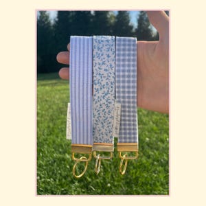 May include: Three fabric wristlet keychains with gold-tone metal hardware and circular rings. The left features light blue vertical stripes, the center has a blue floral pattern on white, and the right displays a light blue and white gingham check.