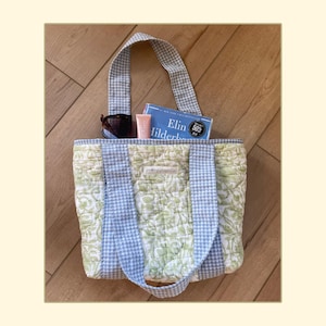 May include: A quilted tote bag featuring a light green and white floral pattern with light blue and white gingham check straps and trim. Inside the bag are a pair of dark sunglasses, a small pink tube, and a book titled Elin Hilderbrand.