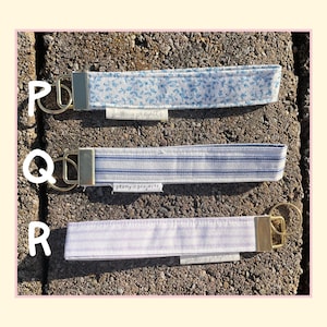 May include: Three fabric wristlet keychains with gold-tone metal hardware and split rings. Top keychain P features a blue floral pattern. Middle keychain Q has blue and white stripes. Bottom keychain R has light purple stripes. Each has a small tag.