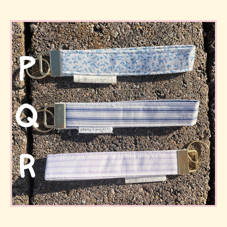 May include: Three fabric wristlet keychains with gold-tone metal hardware and split rings. Top keychain P features a blue floral pattern. Middle keychain Q has blue and white stripes. Bottom keychain R has light purple stripes. Each has a small tag.