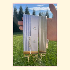 May include: Three fabric wristlet keychains with gold-tone hardware and split rings. One features blue and white vertical stripes, one is white with small floral motifs, and one is sage green with thin white vertical stripes. Each has a small tag.