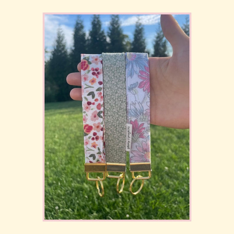 May include: Three fabric wristlet keychains with gold-tone metal hardware and split rings. The left features a pink and orange floral print, the center has a small green and white pattern, and the right displays a blue and pink floral design.