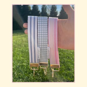 May include: Three fabric wristlet keychains with gold-tone metal hardware. One features blue and white vertical stripes, one has a grey and white gingham check pattern, and one displays pink and white vertical stripes. Each has a Peony Projects label.