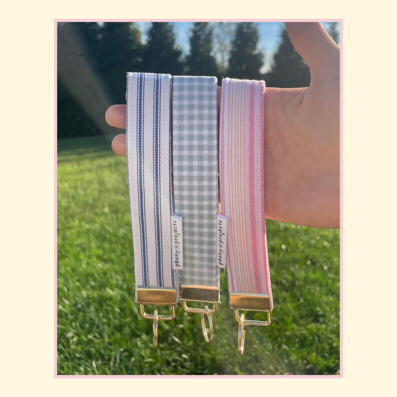 May include: Three fabric wristlet keychains with gold-tone metal hardware. One features blue and white vertical stripes, one has a grey and white gingham check pattern, and one displays pink and white vertical stripes. Each has a Peony Projects label.