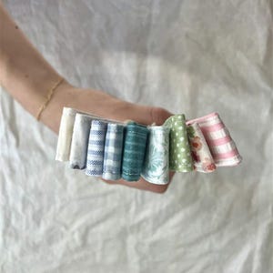 A collection of small, colorful fabric scraps held in a hand. The fabrics display various patterns, including gingham, polka dots, and floral designs, in shades of blue, green, pink, and white. The fabrics are arranged in a fan-like shape.