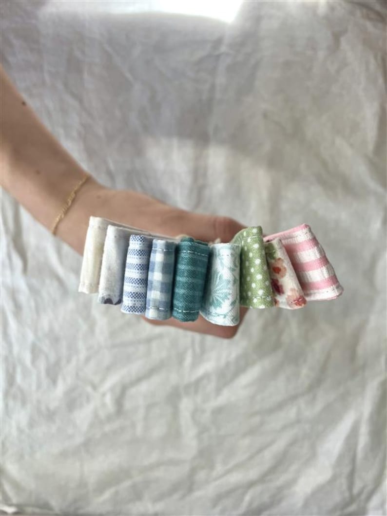 A collection of small, colorful fabric scraps held in a hand. The fabrics display various patterns, including gingham, polka dots, and floral designs, in shades of blue, green, pink, and white. The fabrics are arranged in a fan-like shape.