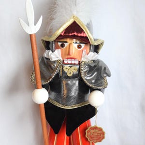 May include: Wooden nutcracker figurine of Captain Guido of the Swiss Guards, wearing a silver and black uniform with red and white striped pants. The figurine holds a wooden spear and has a white feather plume.