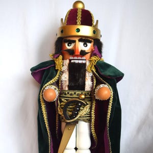 May include: A wooden nutcracker figure wearing a gold crown with green gems, a burgundy velvet hat, and a dark green cape with purple lining. The figure has a black beard, white trousers, black boots with gold laces, and holds a gold sword.