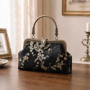 Puede incluir: Un bolso de mano negro con un marco de metal dorado y un asa arqueada a juego. La tela está decorada con un patrón repetitivo de pequeñas ramas de cerezo en flor blancas y doradas. El bolso se cierra con un mecanismo de cierre metálico central.
