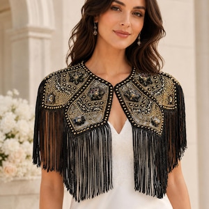 Puede incluir: Una chaqueta bolero negra con intrincados abalorios dorados y negros, lentejuelas y bordados florales sobre una base de malla transparente. La prenda está rematada con largos flecos negros colgantes en los bordes y un escote abierto redondeado.