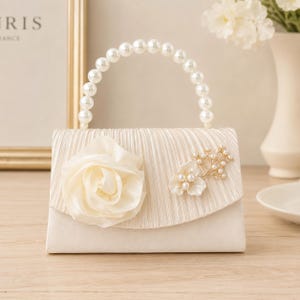 Puede incluir: Un bolso de mano color crema con solapa plisada, asa superior de perlas artificiales blancas, una gran rosa de tela a la izquierda y un adorno floral dorado con pequeñas perlas en el lado derecho de la solapa.