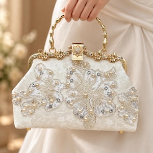 Puede incluir: Un bolso de mano blanco con intrincados bordados florales, lentejuelas plateadas y perlas de color crema. El bolso cuenta con un marco de metal dorado, un cierre decorativo con motivo floral y un asa dorada con estilo de bambú.