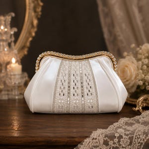 Puede incluir: Un bolso de mano de satén blanco con un marco de metal dorado incrustado con cristales transparentes. El panel central está decorado con filas verticales de pequeñas cuentas blancas y detalles plateados rectangulares, con un cierre superior curvo.